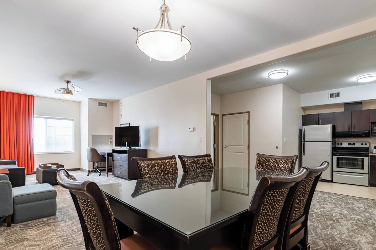A modern dining area with a glass table, four upholstered chairs, a ceiling fan, and a kitchen in the background.