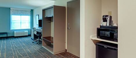 Een kleine hotelkamer met een kitchenette, een bureau en een raam met zonwering.
