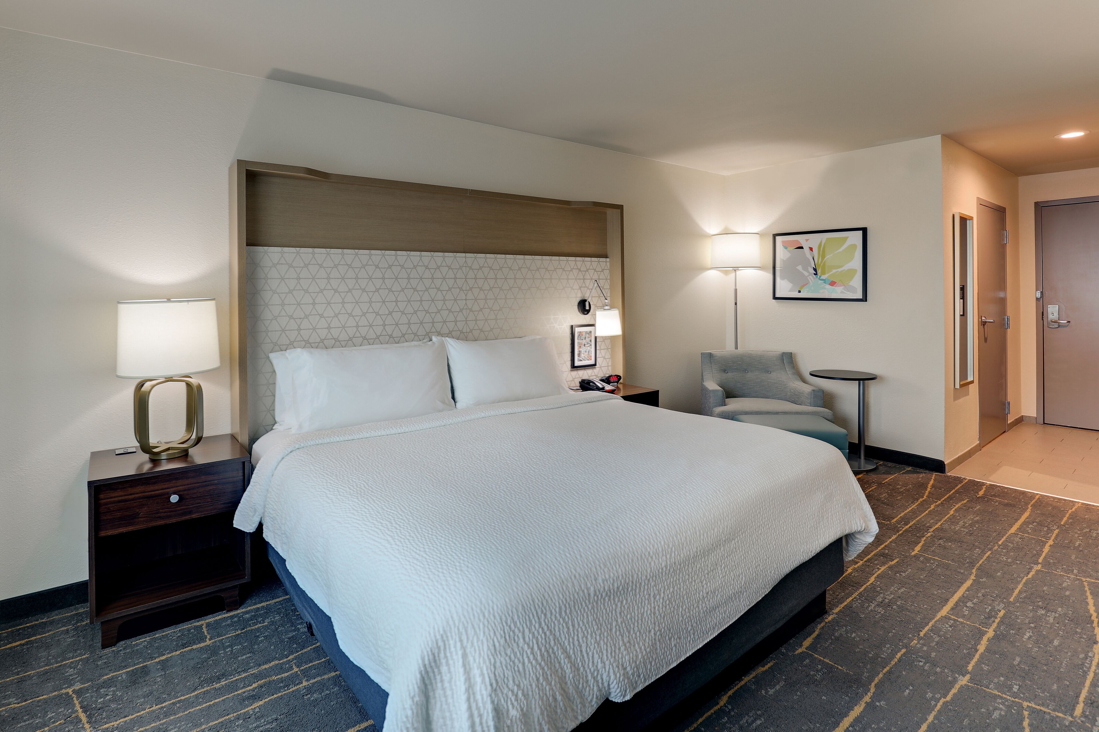 A hotel room with a large bed, bedside tables, a chair, and a lamp.