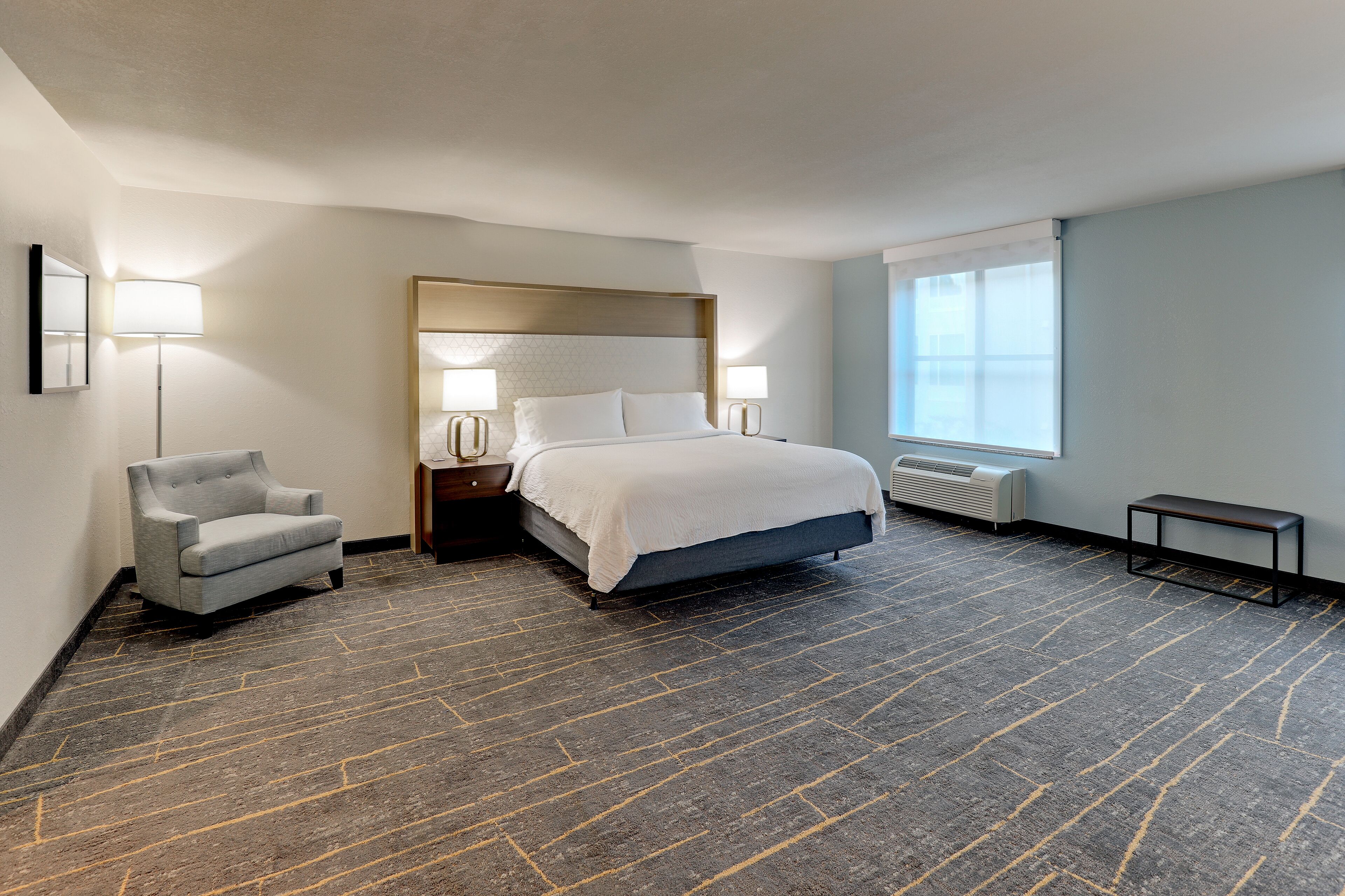 A hotel room with a bed, bedside lamps, a chair, a small table, and a window.