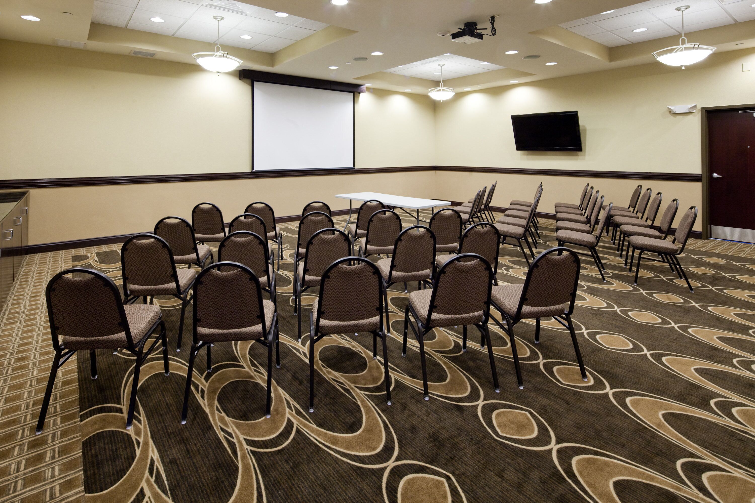 A conference room with rows of chairs, a projector screen, and a flat-screen TV on the wall.