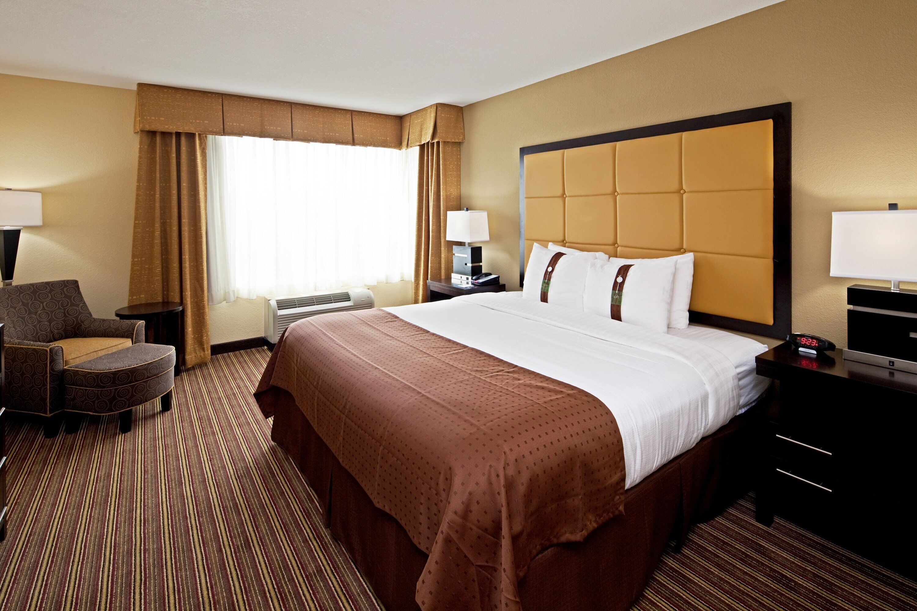 Una habitación de hotel con una cama grande, un escritorio con lámpara, una silla, una ventana con cortinas y un reloj de pared.