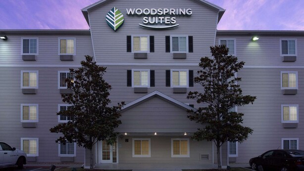 Un hôtel à plusieurs étages, avec une enseigne indiquant « Woodspring Suites ». Des arbres se trouvent devant l’immeuble, et un parking est aménagé avec des voitures.