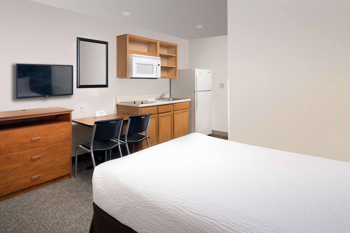A hotel room with a bed, a desk with a chair, a microwave, a refrigerator, a TV, and a wooden dresser.