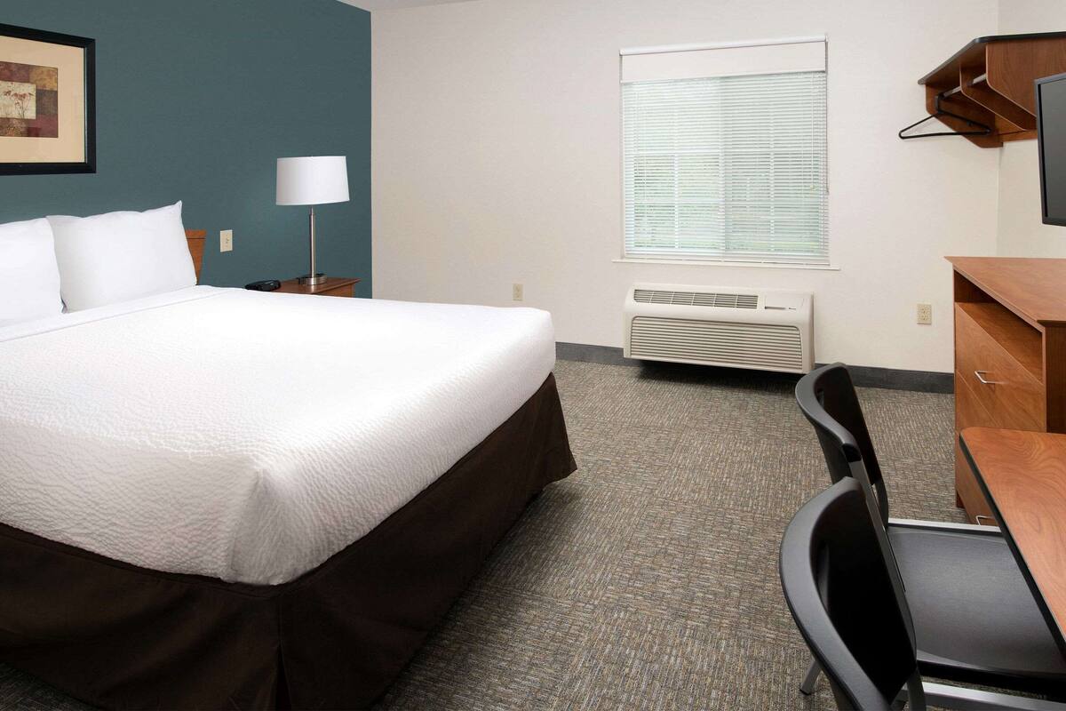 A hotel room with a large bed, a desk, a chair, a lamp, a window with blinds, and an air conditioning unit.