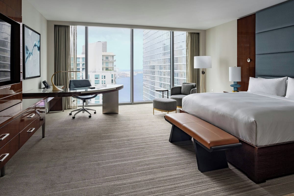 A hotel room with a large bed, a desk with a chair, a small table, and a bench.