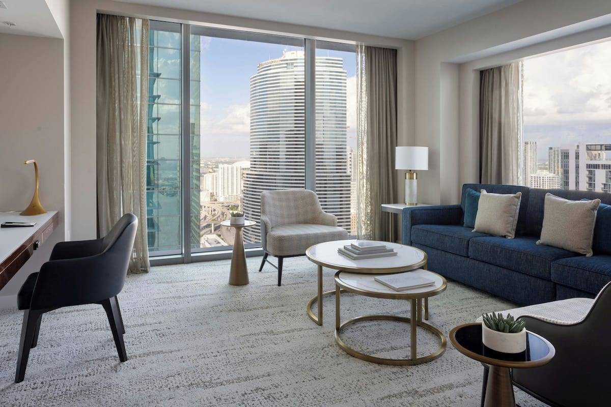 A modern hotel room with a large window, a sofa, a chair, a small table, and a desk.