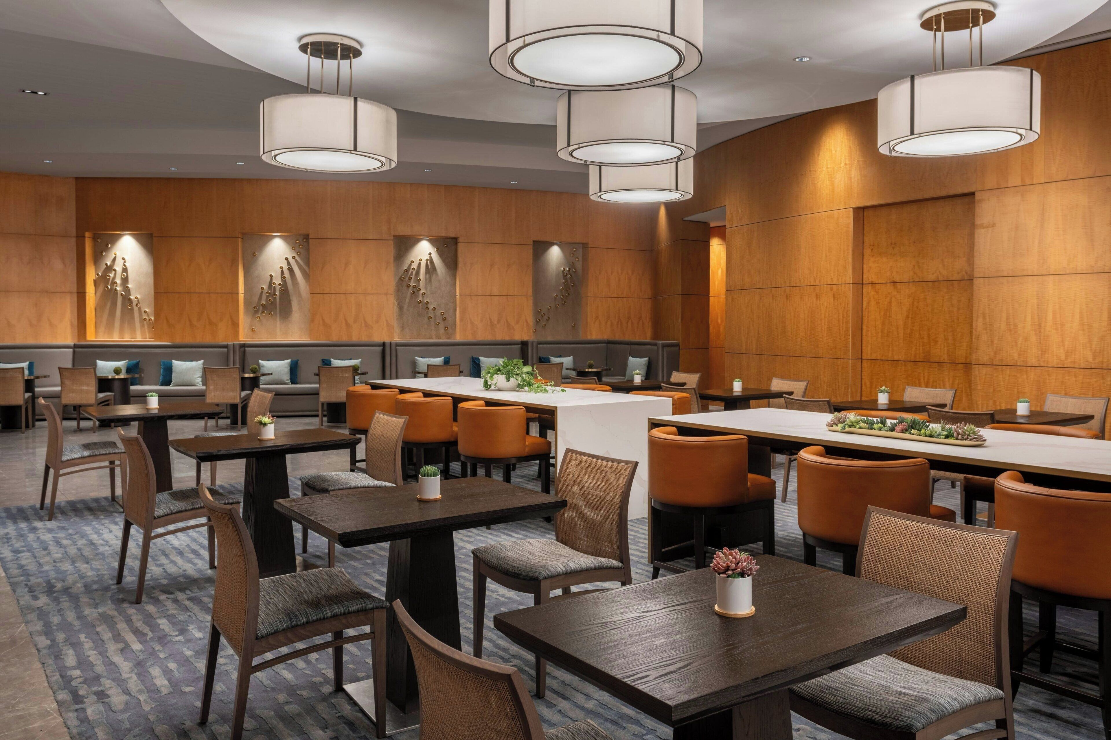 A hotel dining area with multiple tables and chairs, wooden panel walls, and pendant lighting.
