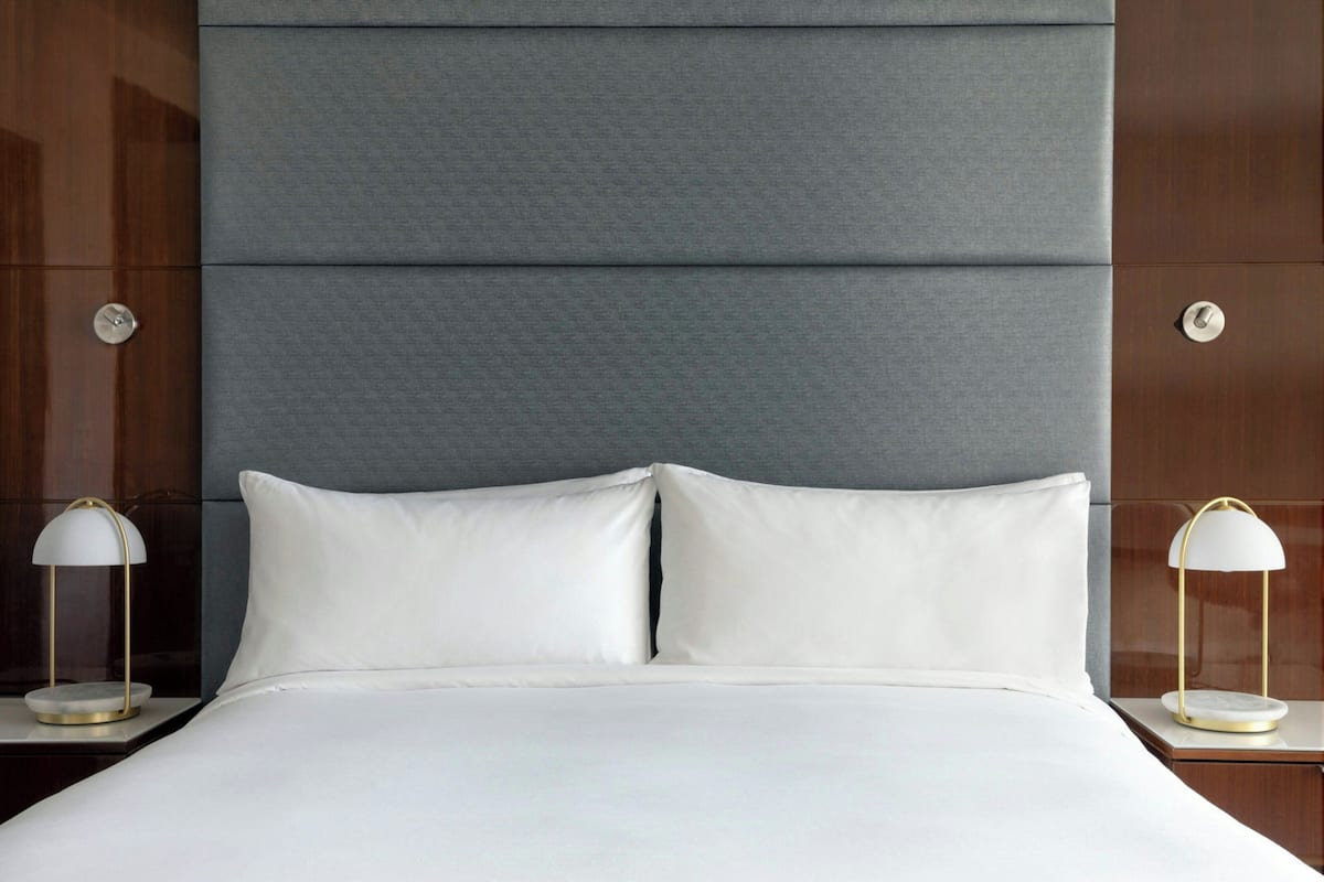 A neatly made bed with white linens, flanked by two bedside tables with lamps, set against a wall with a textured panel and a wooden finish.