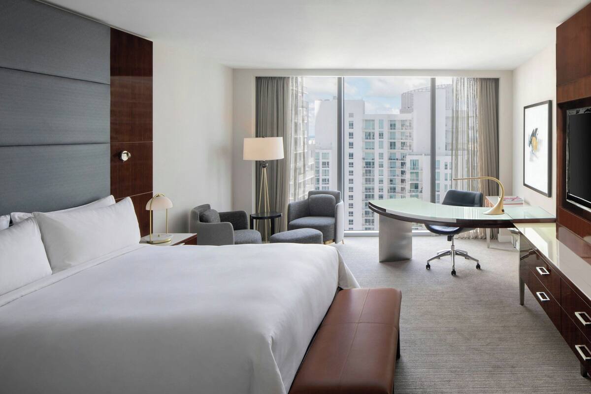 A modern hotel room with a large bed, a desk, a chair, and a view of the cityscape.