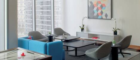A modern living room with a blue sofa, two armchairs, a coffee table, and a foosball table. There is a large window with a city view, a geometric painting on the wall, and a small plant on the coffee table.