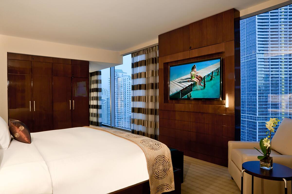 A hotel room with a large bed, a TV mounted on the wall, a sofa, and a view of a cityscape through large windows.