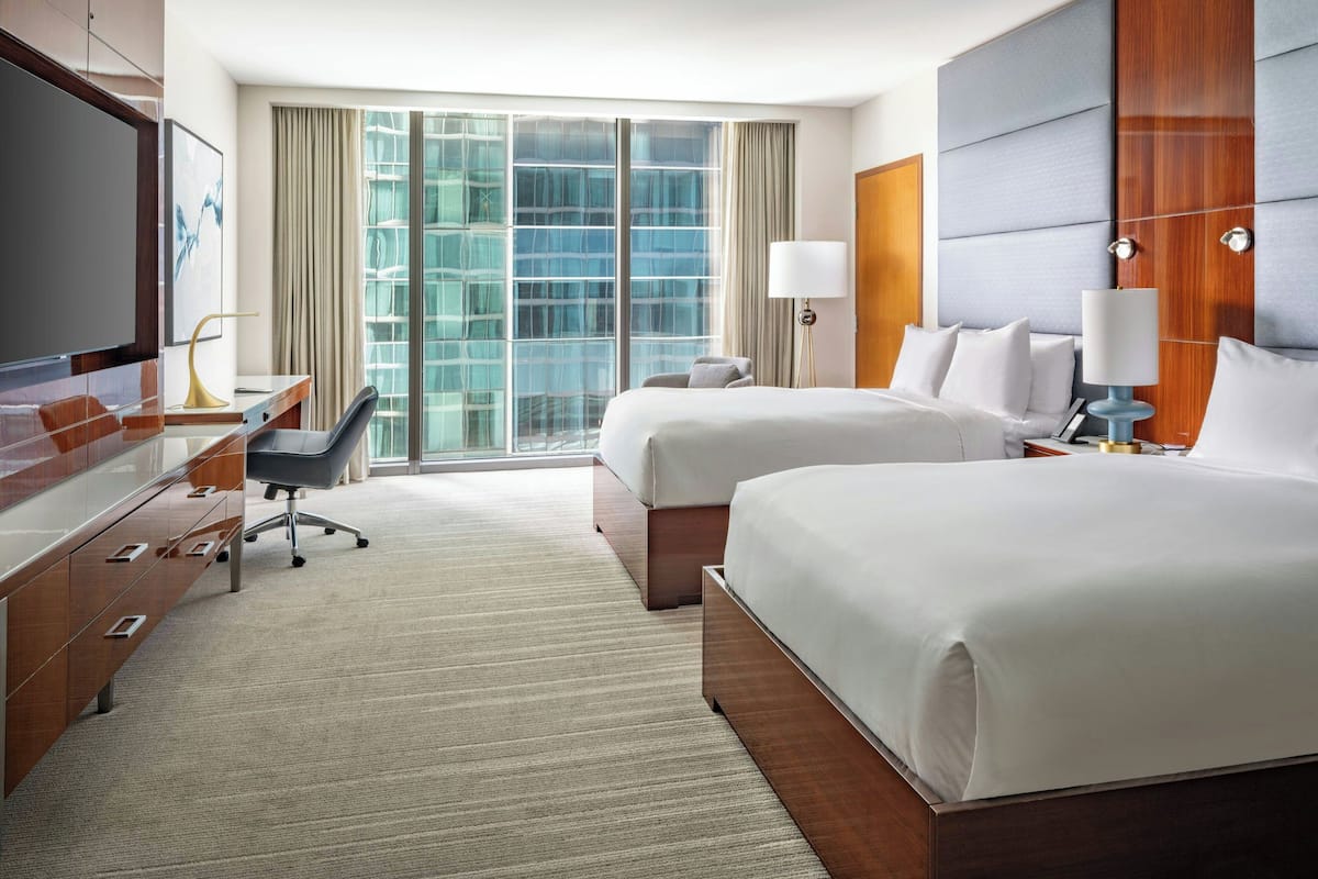 A hotel room with two beds, a desk with a chair, a TV, and a large window with curtains.