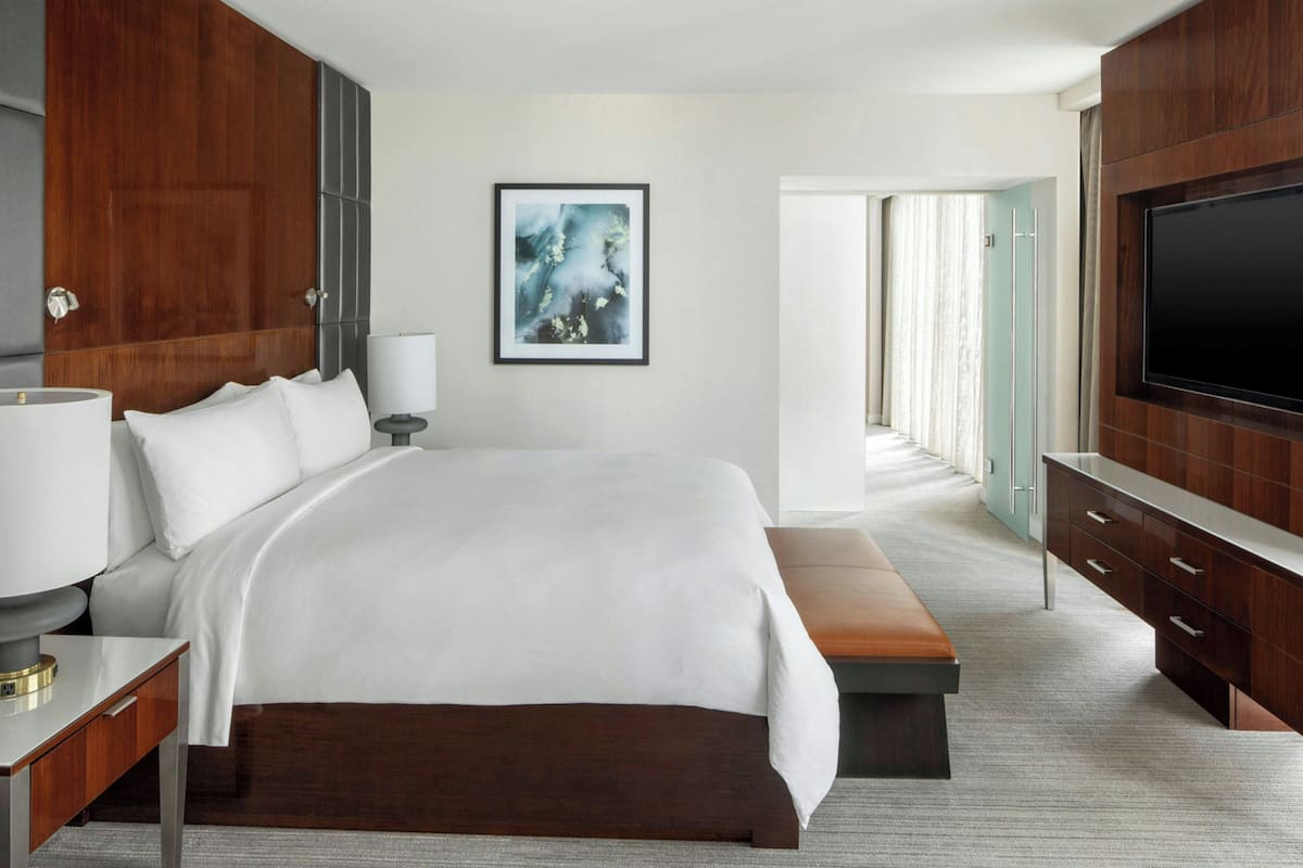A modern hotel room with a large bed, a bedside table with a lamp, a bench, a TV, and a framed picture on the wall.
