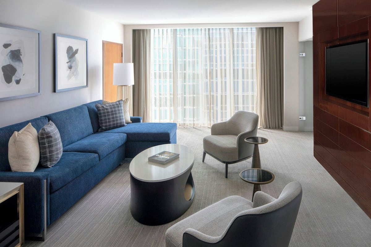 A modern hotel room with a blue sofa, a grey armchair, a round coffee table, and a flat-screen TV mounted on a wooden panel.