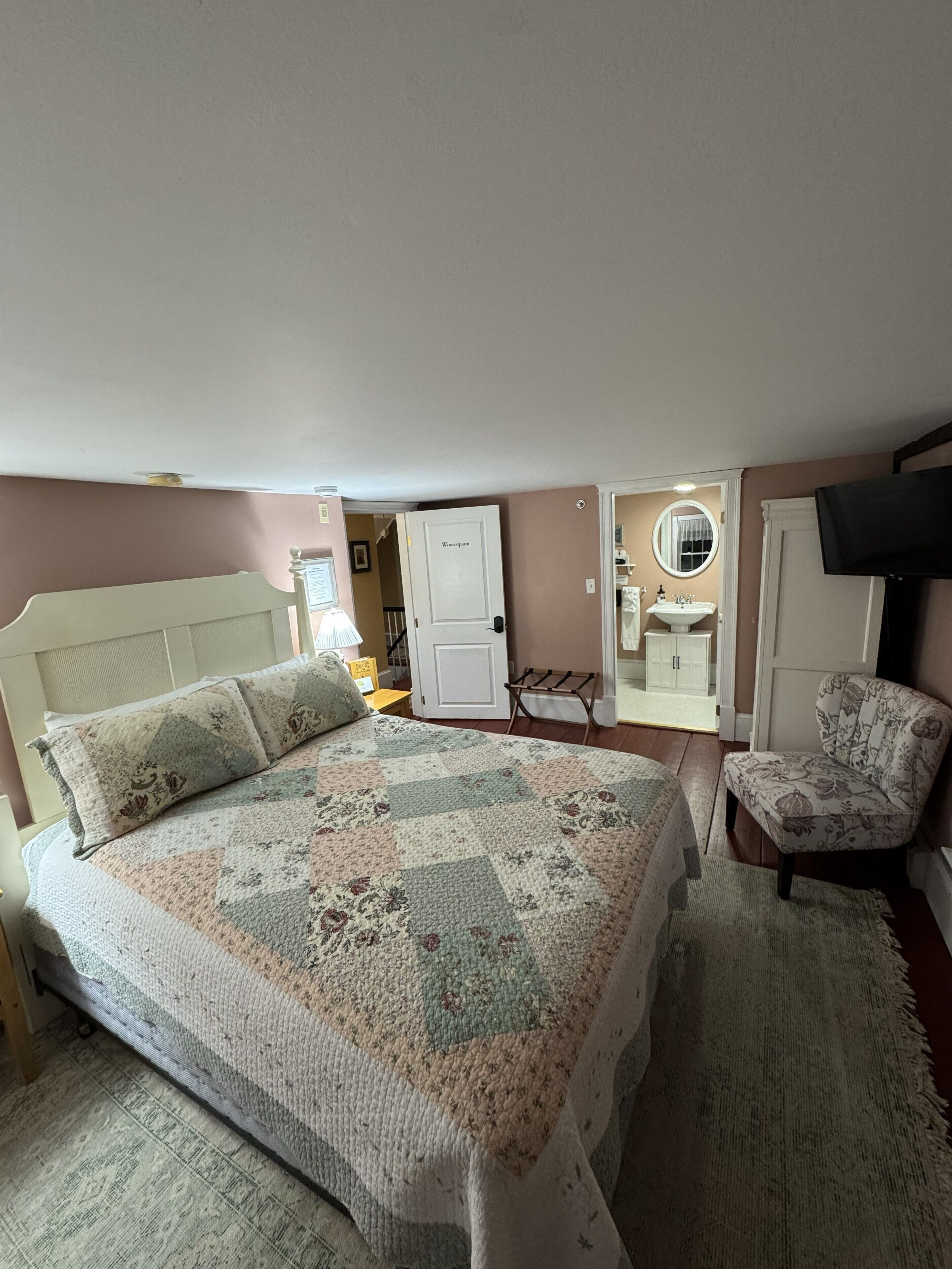 Une chambre à coucher avec un lit, une chaise, un téléviseur, une porte et une salle de bain visible à travers une porte ouverte.