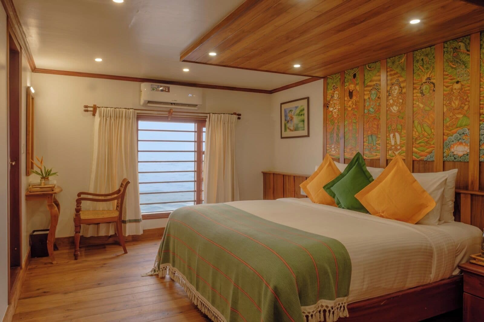 A bedroom with a wooden ceiling, a bed with green and yellow pillows, a wooden chair, a window with curtains, and a wall with a colorful tapestry.