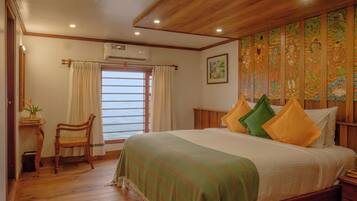 A bedroom with a wooden ceiling, a bed with green and yellow pillows, a wooden chair, a window with curtains, and a wall with a colorful tapestry.
