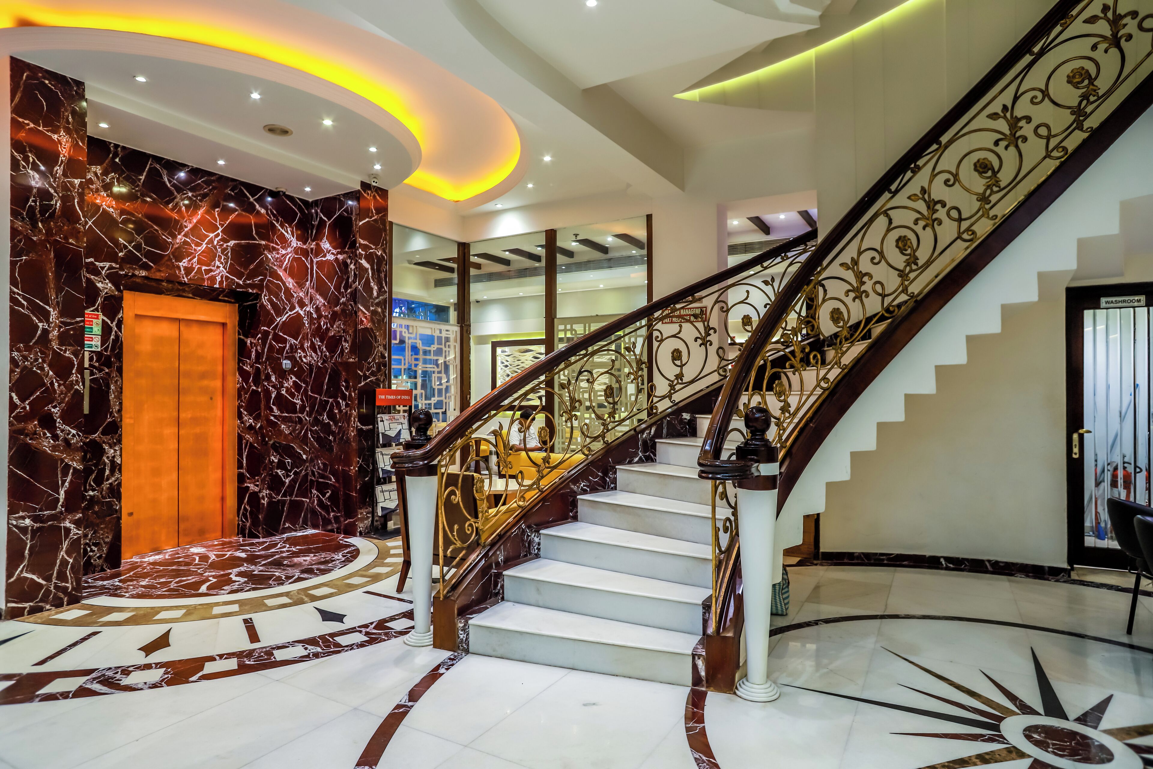 Una gran escalera con suelo de mármol y barandillas ornamentales, que conduce a un ascensor y una recepción con pared de mármol y suelo de diseño circular.