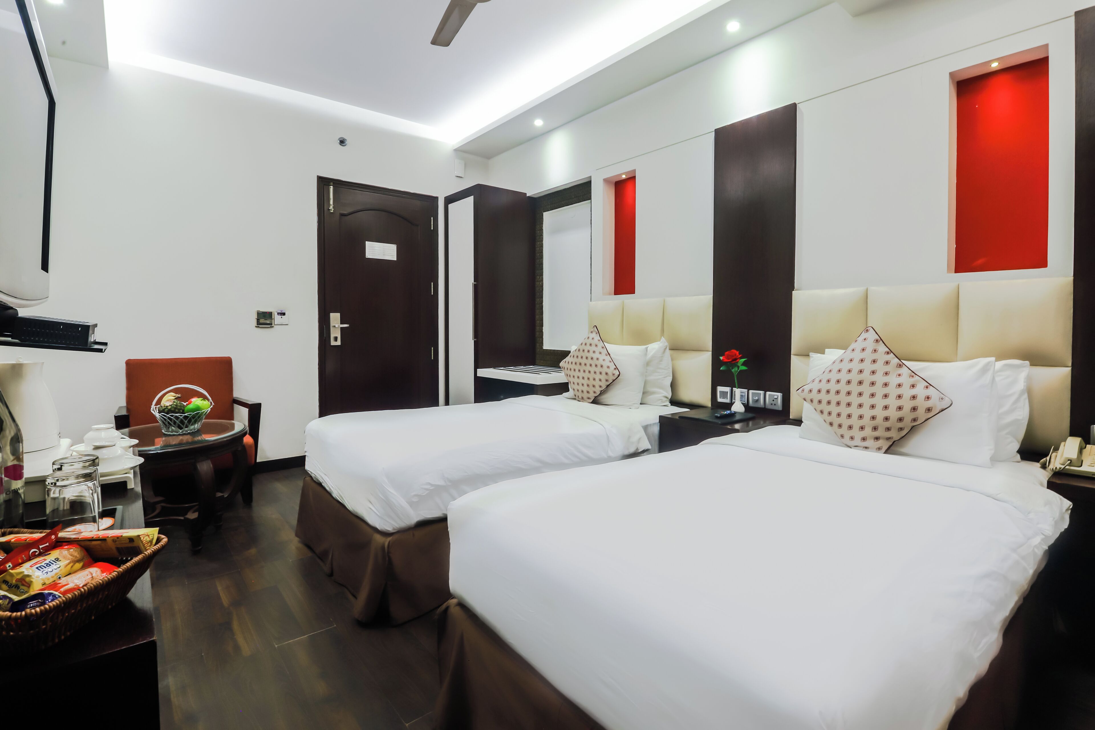 A hotel room with two beds, a desk with a chair, a small table with a basket of snacks, and a red accent wall.
