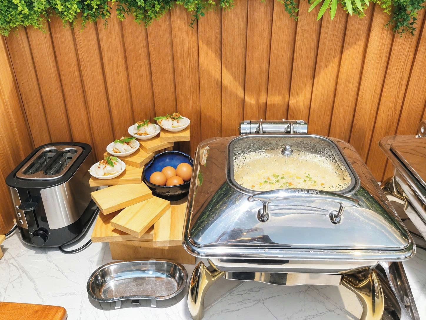 A kitchen area with a toaster, a food station with plates of food, a bowl of eggs, and a large metal container.