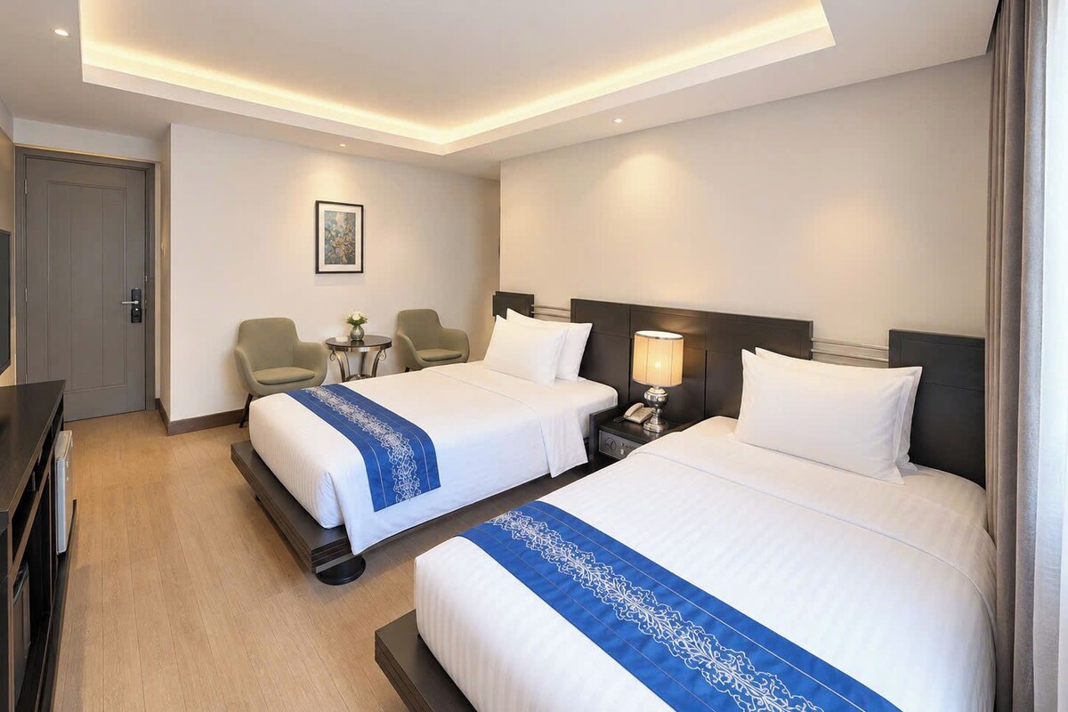 Ein Hotelzimmer mit zwei Betten, einem Schreibtisch, einem Stuhl, einem Nachttisch und einem kleinen Tisch mit einer Lampe.