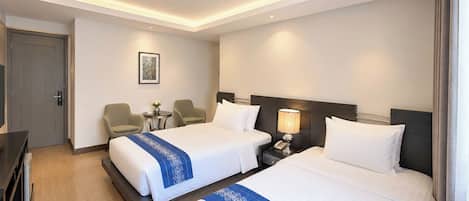 A hotel room with two beds, a desk, a chair, a nightstand, and a small table with a lamp.