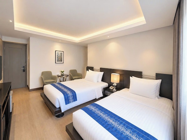 A hotel room with two beds, a desk, a chair, a nightstand, and a small table with a lamp.