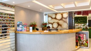 A reception desk with a curved design, a wall with a textured pattern, and a shelf with various items including books and flowers. - A&EM - The Petit Hotel (Ho Chi Minh City)