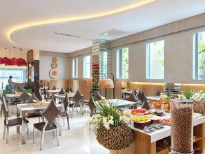 Ein moderner Essbereich mit mehreren Tischen und Stühlen, ein Buffet mit verschiedenen Speisen und große Fenster, die für natürliches Licht sorgen.