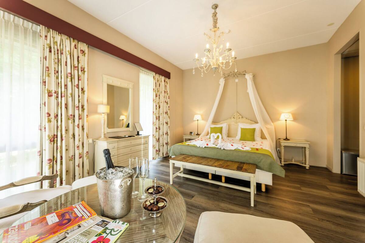 Una habitación de hotel con una cama con dosel, una araña, una mesa de comedor con una jarra de champán y una bandeja de desayuno.