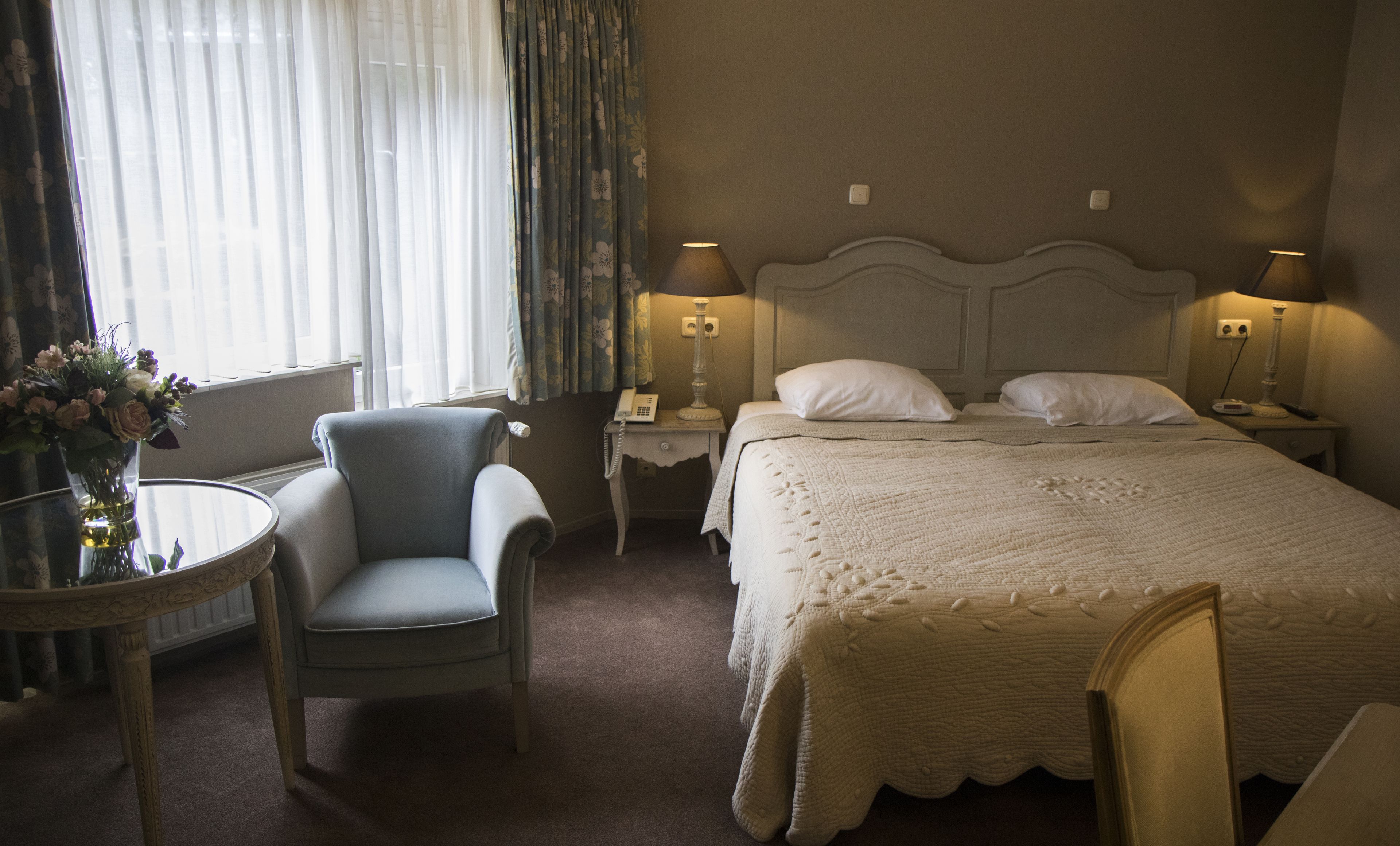 A hotel room with a bed, two bedside lamps, a chair, a table with a vase of flowers, and a window with curtains.