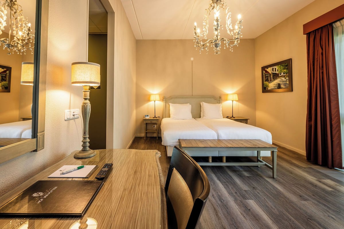 Una habitación de hotel con cama, mesitas de noche, un escritorio con silla, un espejo y una araña.