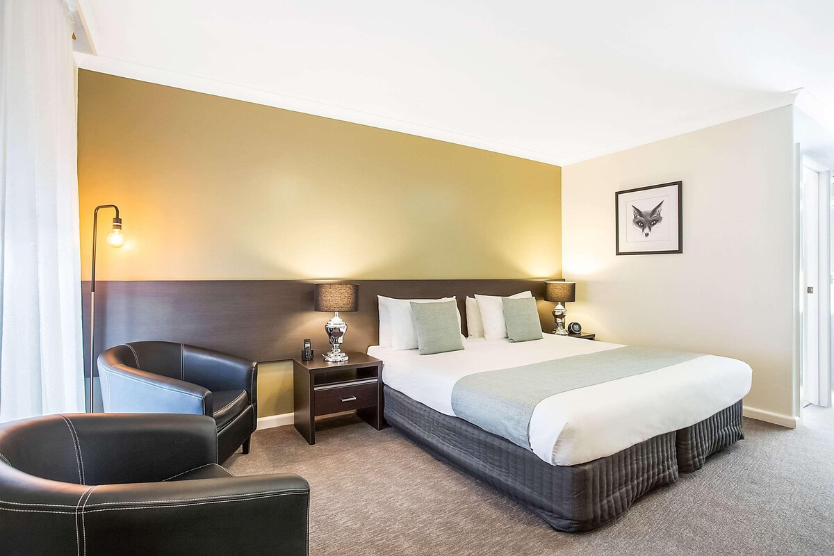 Habitación de hotel con una cama grande, una silla, una mesita de noche y una lámpara fijada a la pared.