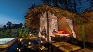 Zona de estar al aire libre con techo de paja, piscina rodeada de árboles e iluminación nocturna.