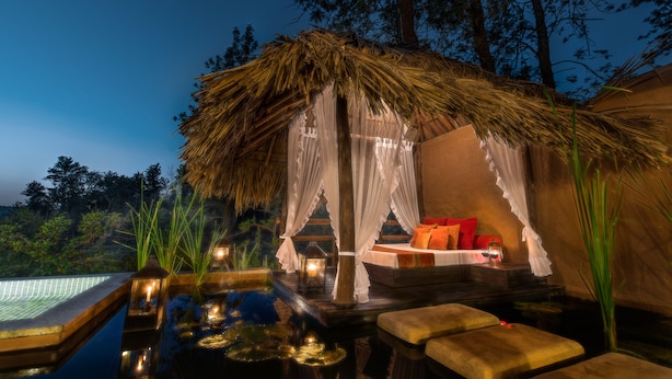 A thatched-roof outdoor seating area with a pool, surrounded by trees and night lighting. - Evolve Back, Coorg (Virajpet)