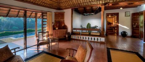 A spacious room with a high ceiling, wooden furniture, and large windows offering a view of a lush garden.