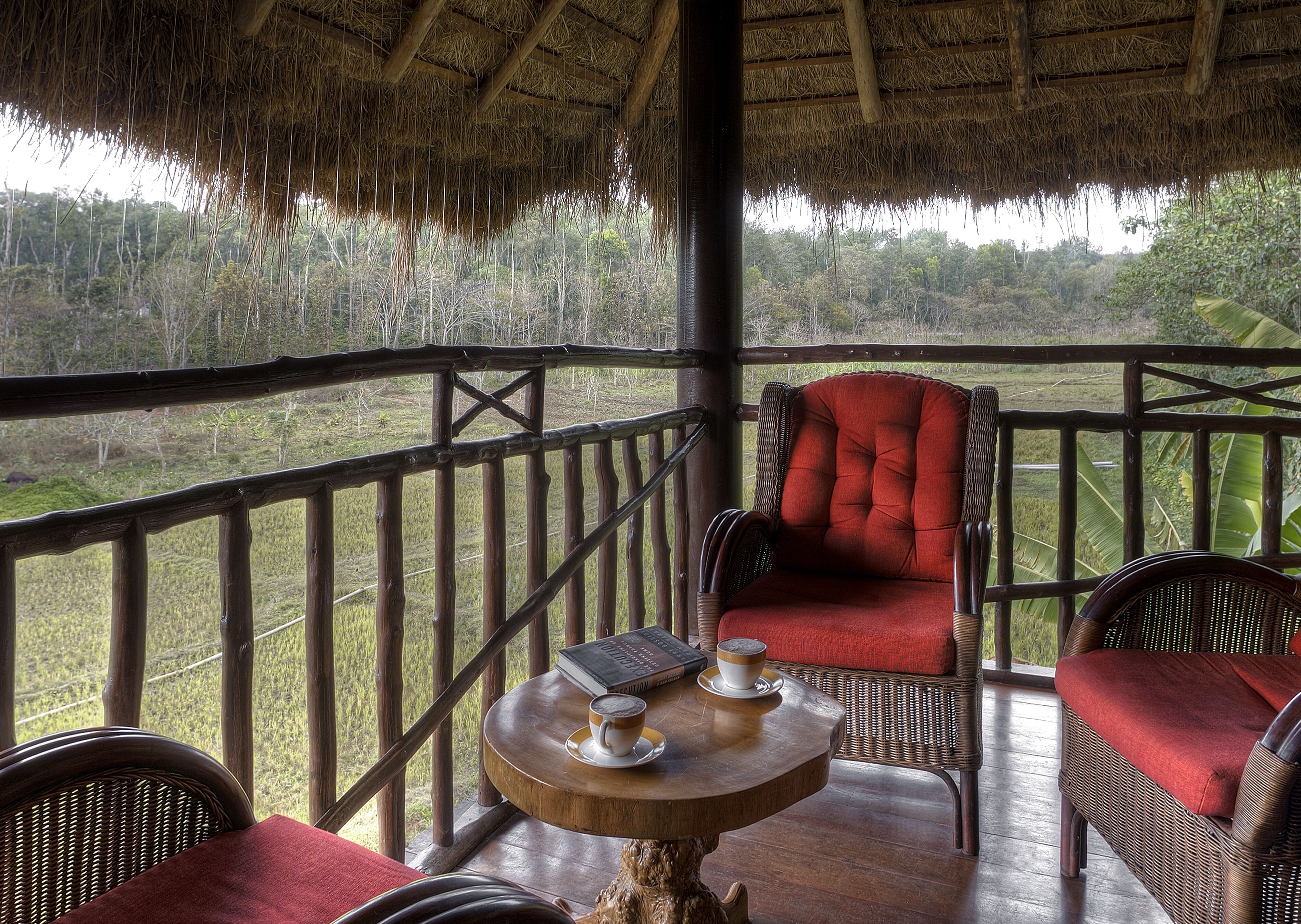 Terraza de madera con mobiliario de mimbre y una silla con cojín rojo, con vistas a un paisaje exuberante.
