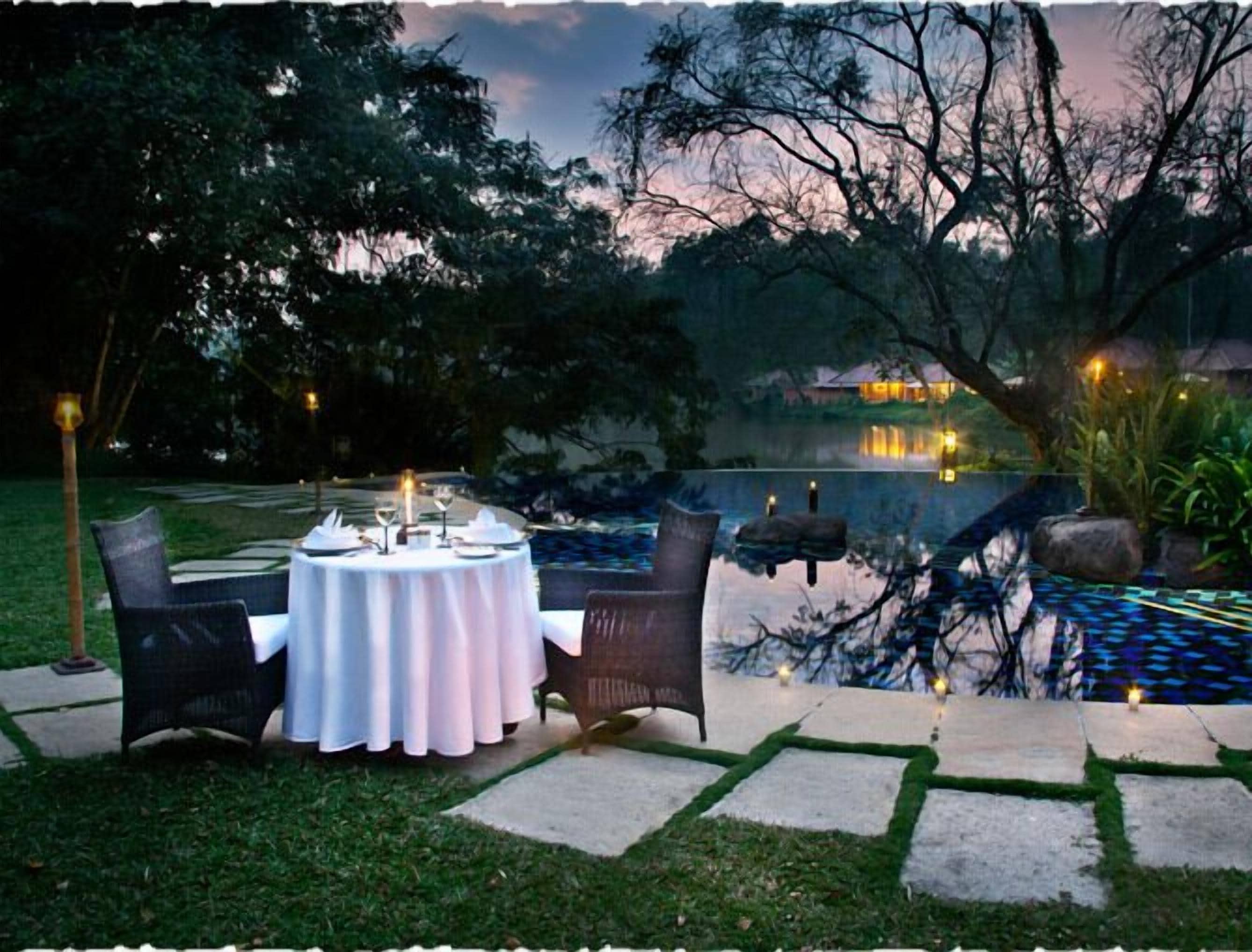 An outdoor dining area with a round table set for two, surrounded by chairs, near a pool with a view of a house in the background.
