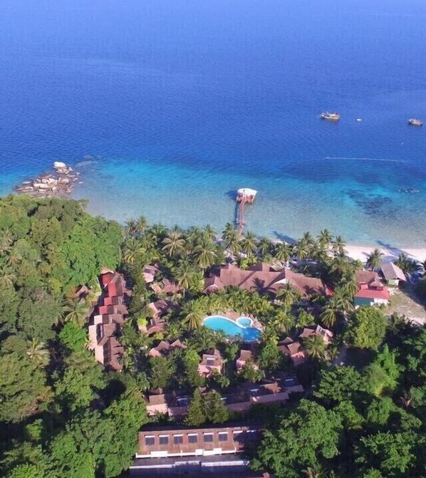 An aerial view of a resort with a swimming pool, palm trees, and a beach.