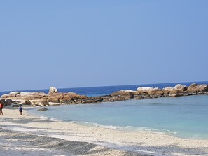 A beach with white pebbles, clear blue water, and large rocks.