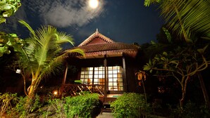 A traditional thatched-roof hut with a large window, surrounded by lush greenery and a pathway leading to it.