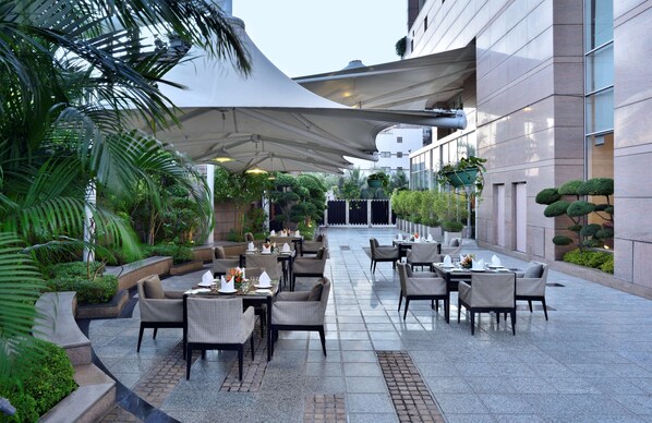 A spacious outdoor dining area with multiple tables and chairs, surrounded by lush greenery and a modern building facade.