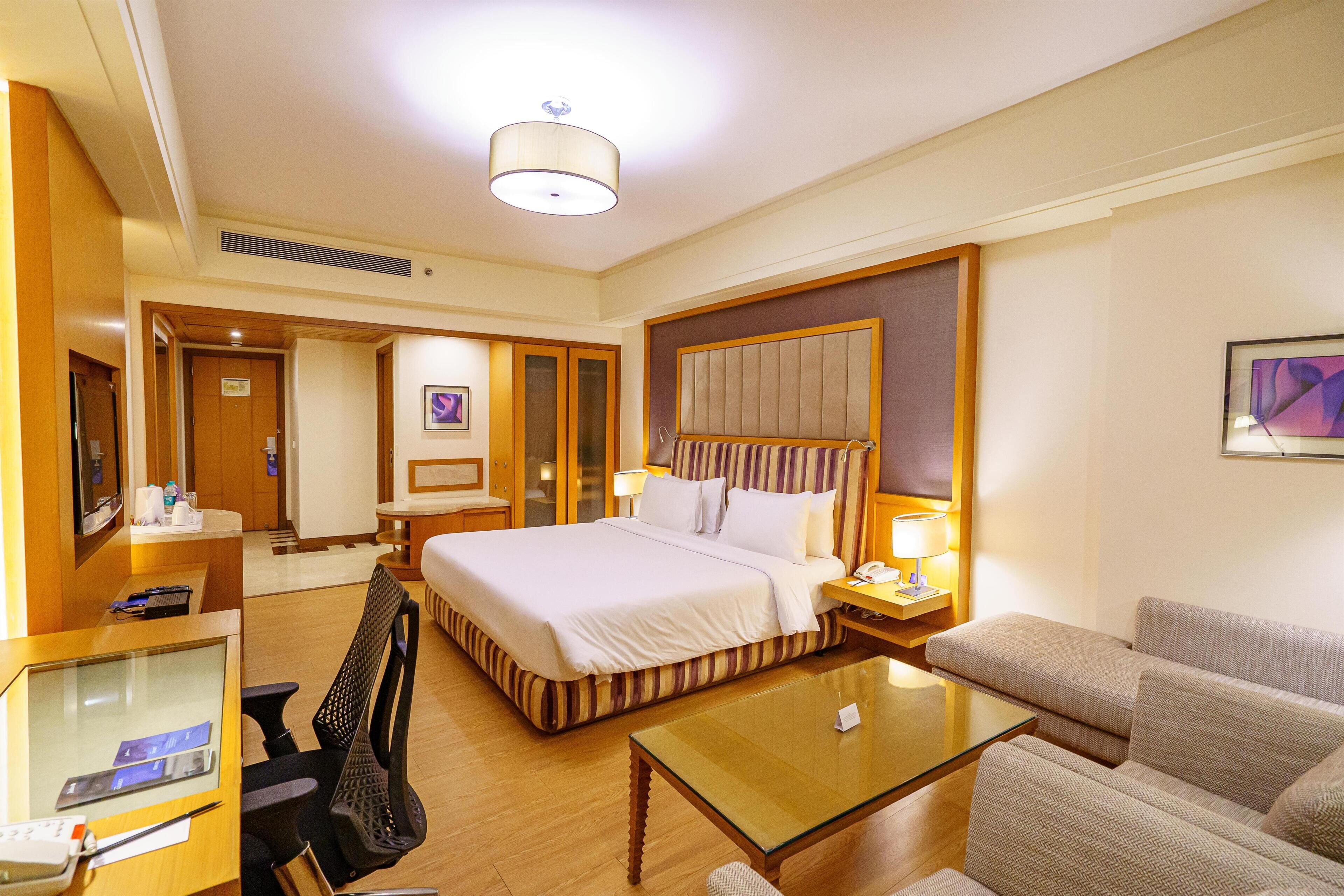 A hotel room with a bed, a sofa, a desk with a computer, a chair, and a bedside table with a lamp.