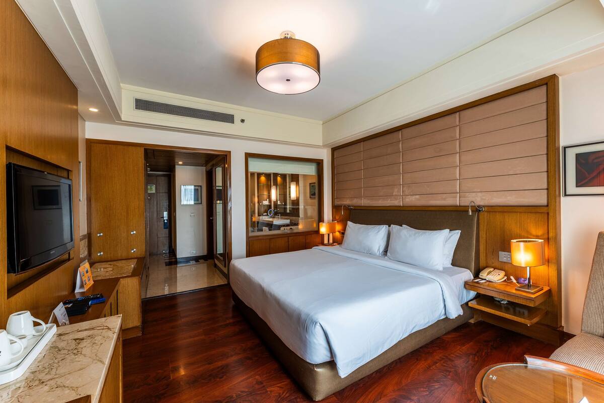 Habitación de hotel con una cama grande, un escritorio con televisor de pantalla plana, una mesita de noche con lámpara y un espejo sobre la cama.