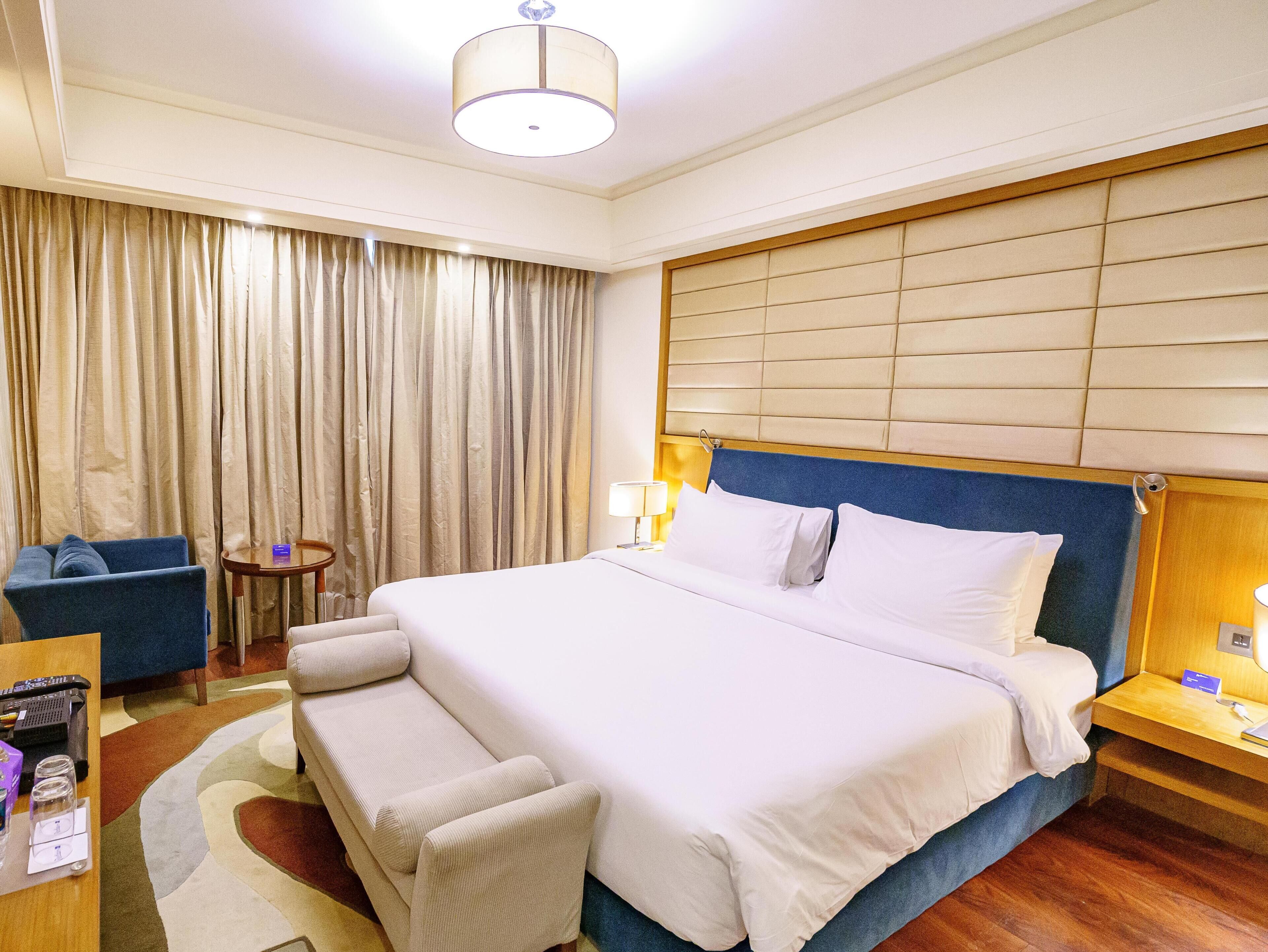 A hotel room with a large bed, a blue armchair, a small table, and a desk with a lamp.