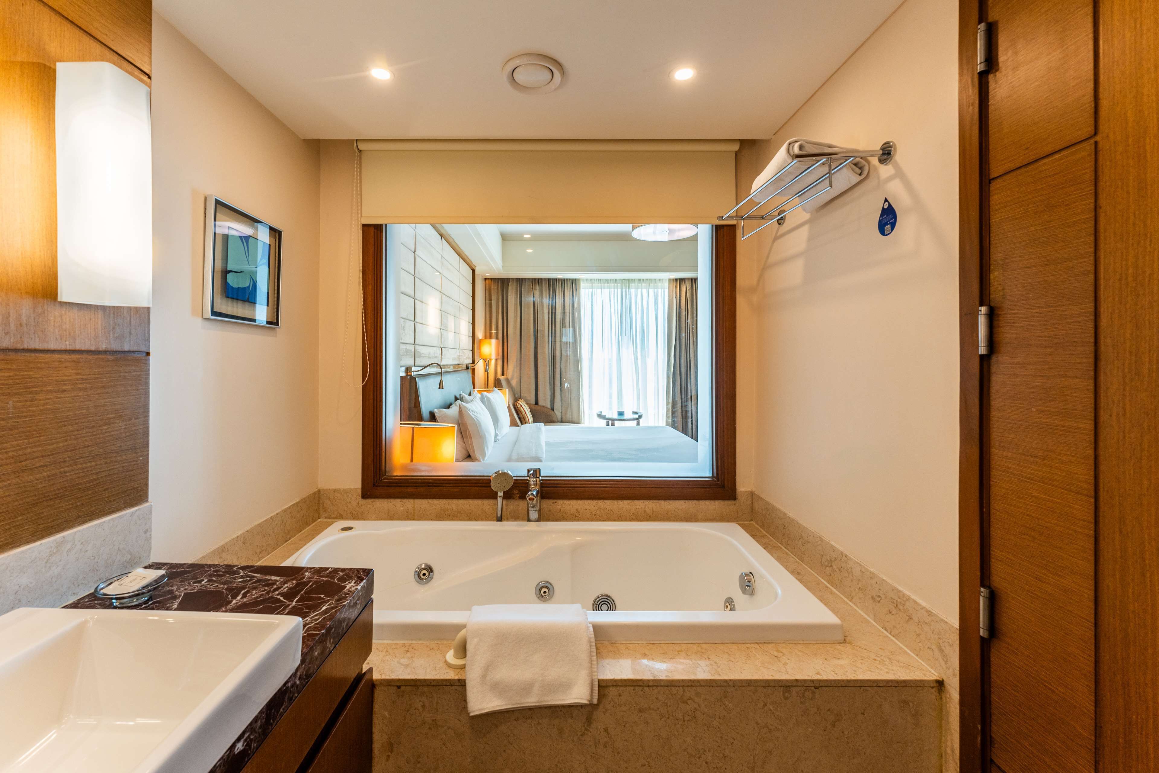 A modern bathroom with a large bathtub, a separate sink, and a mirror reflecting a bedroom with a bed and curtains.