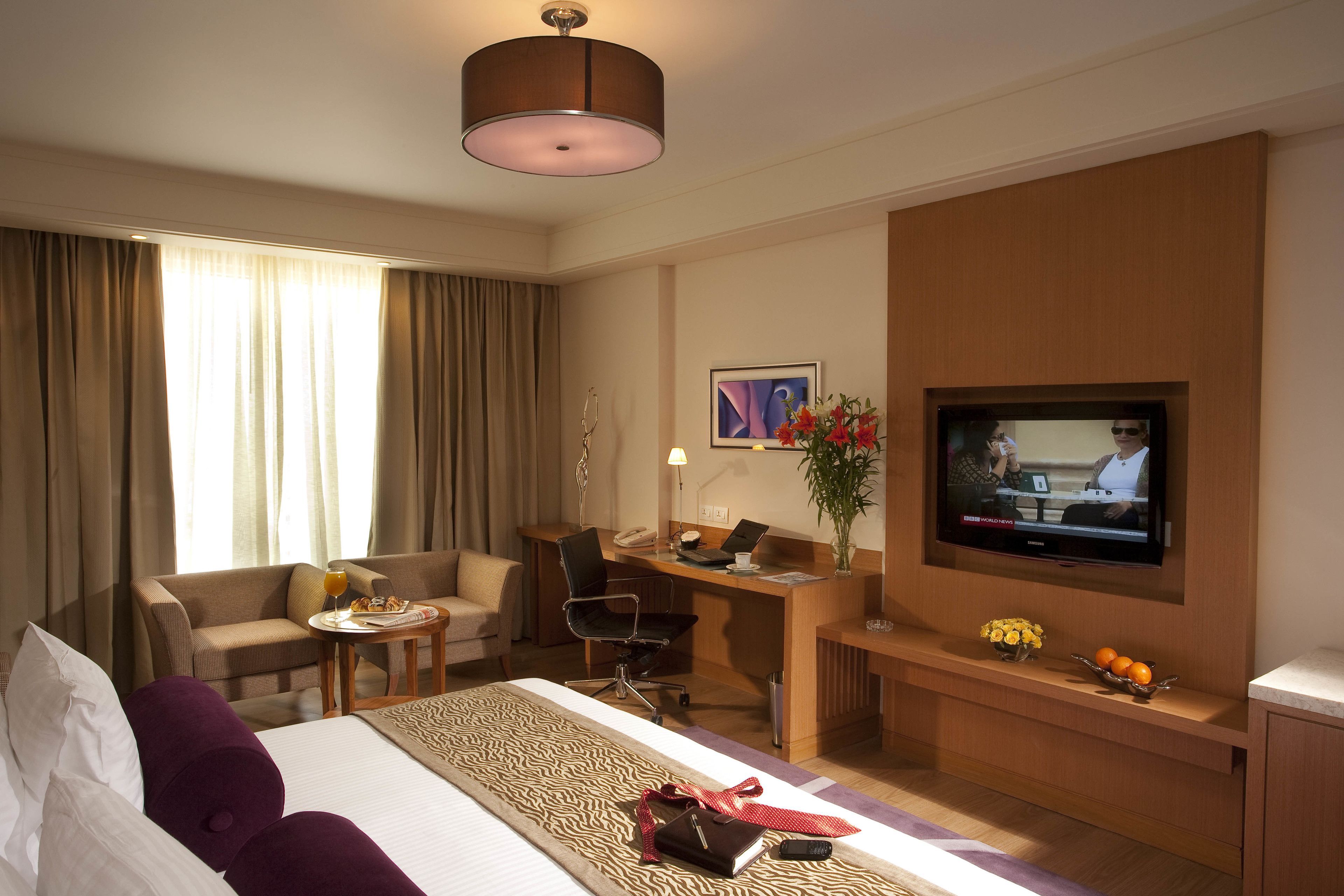 A hotel room with a bed, a desk, a television, a sofa, and a small table with a plant and fruit.