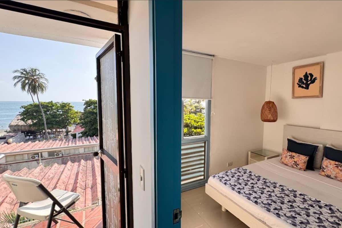 A bedroom with a bed, pillows, a nightstand, and a view of the sea through a balcony.
