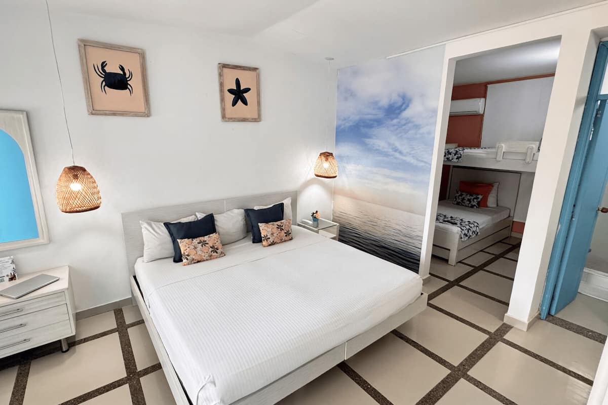 A modern bedroom with a large bed, a bedside table, a dresser, and a mirror. There are two wall art pieces, a ceiling mural, and a door leading to another room.